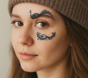 En ung person med nesering og brun strikkelue bærer Blå og brune øyesminketatoveringer, med kunstneriske blå påfuglformede motiver over og under høyre øye. De ser rolig inn i kameraet med et nøytralt uttrykk.