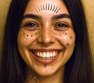 En smilende kvinne med langt mørkt hår og fregner bærer Gullstjerne-ansiktstatoveringer-dekorative klistremerker med stjerne og strek - med en piercing under underleppen, og står mot en beige bakgrunn.