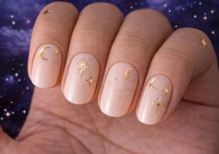 En hand med ljusa nudefärgade nagellack har himmelska mönster i guld, inklusive månar och stjärnor, mot en bakgrund av en stjärnklar natthimmel.
