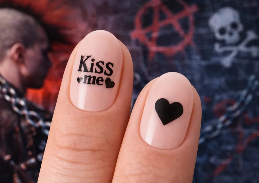 Dos dedos con nail art: una uña dice Bésame con dos corazones pequeños, y la otra tiene un corazón negro. En el fondo borroso se ve a una persona con cresta y símbolos de calavera y anarquía estilo grafiti.