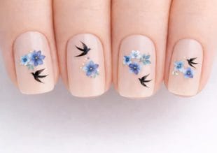Närbild av naglar med nudefärgat nagellack, dekorerade med fina blå blomdekaler och små svarta fågelmotiv på varje nagel.