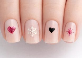 En närbild av fyra naglar med ljusrosa lack, var och en dekorerad med olika motiv: ett rosa glitterhjärta, en vit snöflinga, ett svart hjärta och en rosa glitterstjärnespräckning.