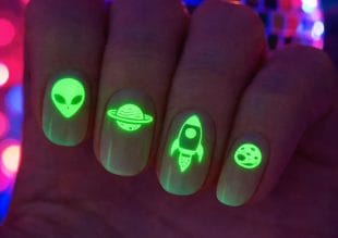 En hand med självlysande nagelkonst med gröna mönster: ett utomjordiskt ansikte, en planet med ringar, en raket och en måne eller asteroid, var och en på en separat nagel. Bakgrunden har färgglada, suddiga lampor.
