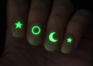 En närbild av fyra fingernaglar, var och en med en lysande neongrön design: en stjärna, en cirkel, en halvmåne och ytterligare en stjärna, allt mot en mörk bakgrund.