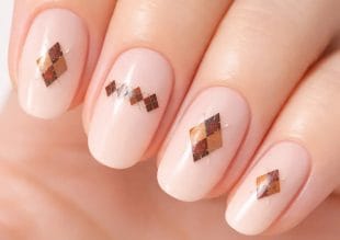 Närbild av en hand med välmanikyrerade naglar, var och en målad i en ljusrosa nyans och dekorerad med bruna och guldfärgade argyle-mönstrade nagelkonstmönster.