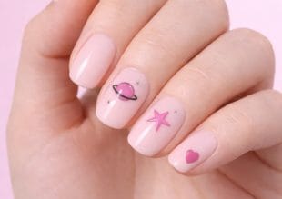 En hand med ljusrosa nagellack med nagelkonst: en rosa planet med en ring, en rosa stjärna och ett rosa hjärta på tre olika naglar. Naglarna är prydligt formade och bakgrunden är ljusrosa.