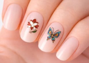 Närbild av en hand med nudefärgat rosa nagellack. En nagel har en röd och vit blomdesign och en annan har en färgglad fjärilsdesign.