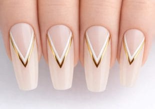 Närbild av fyra fingrar med långa, mandelformade naglar som är målade nakna och dekorerade med chevronmönster i vitt och guld nära nageltopparna.