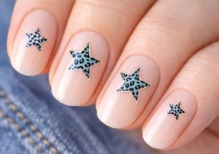 Närbild av fyra naglar målade med ett blekt nudefärgat nagellack, var och en dekorerad med ett blått stjärnformat klistermärke med svart leopardmönster. Bakgrunden innehåller en antydan till denimtyg.