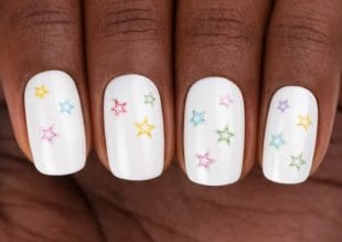 Närbild av en hand med vitt nagellack, där varje nagel är dekorerad med små, färgglada stjärnmotiv i olika nyanser som rosa, gult, grönt, blått och lila.