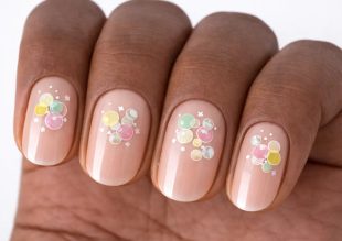 Närbild av fingrar med nudefärgade naglar med färgglada bubbelliknande prickar och små vita accenter som nail art på varje nagel.