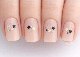 Närbild av naglar målade i en nudefärg och dekorerade med små svarta, guld- och vita stjärnformade nagelkonstverk och små prickar på varje nagel.